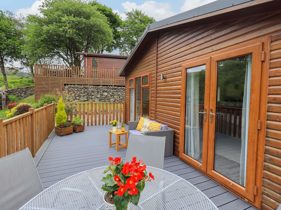 An outdoor deck with wooden railing featuring a round metal table with a red flower pot and chairs beside a wooden cabin with glass doors at Hollytree Lodge in Beckside 1 Limefitt Park near Windermere