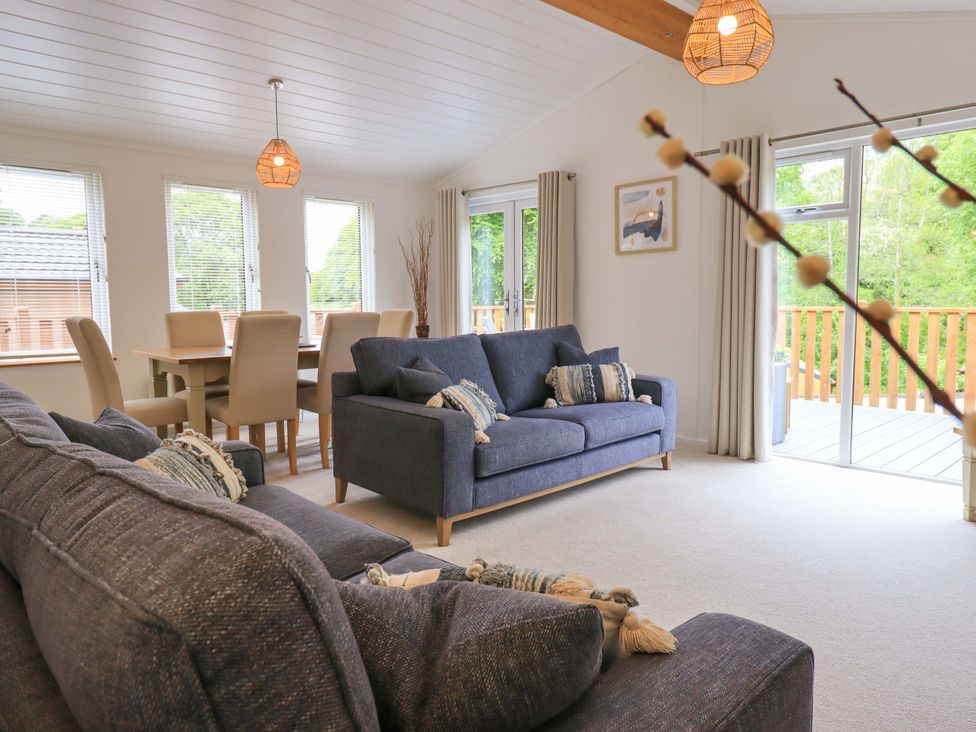 A living room with dark sofas a dining table with beige chairs and large windows at Hollytree Lodge Beckside 1 Limefitt Park near Windermere