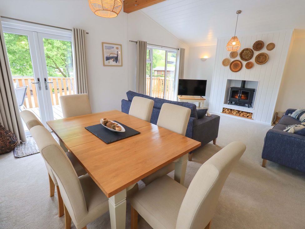 A dining area with a wooden table and beige chairs next to a living room with blue sofas a wood stove and a television at Hollytree Lodge Beckside 1 Limefitt Park near Windermere