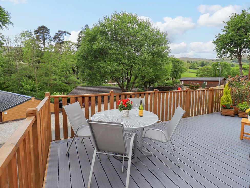 An outdoor deck with a round metal table and four chairs overlooking trees and cabins at Hollytree Lodge Beckside 1 Limefitt Park near Windermere