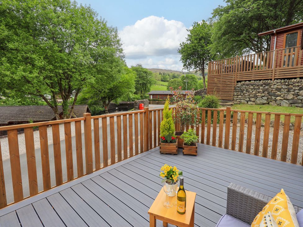 An outdoor deck with wooden railing, potted plants, a small table with wine and glasses, and a chair at Hollytree Lodge Beckside 1 Limefitt Park near Windermere