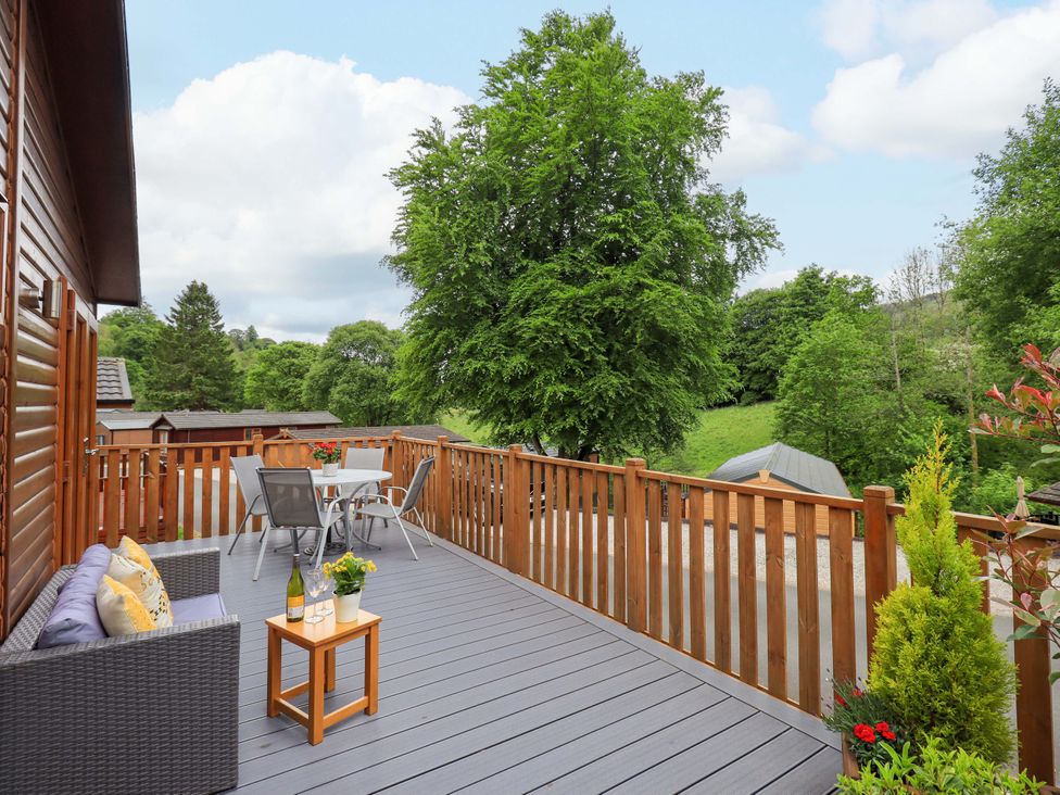 An outdoor wooden deck with gray flooring and wooden railing with a wicker sofa and dining table surrounded by green trees at Hollytree Lodge Beckside 1 Limefitt Park near Windermere