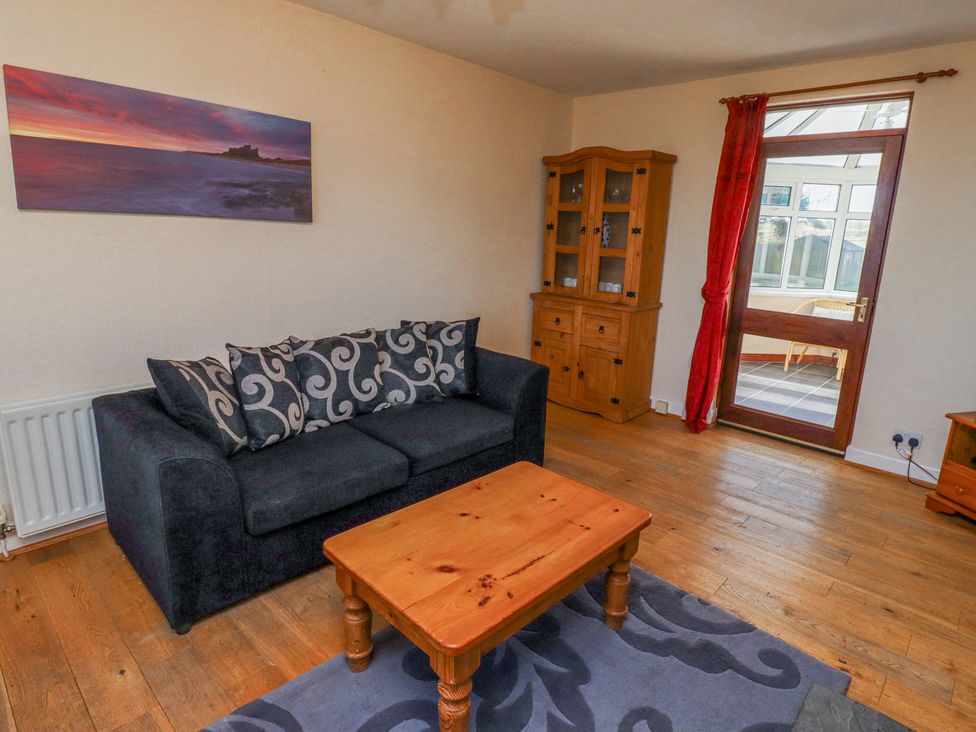 A living room with a black sofa with patterned cushions a wooden coffee table a wooden cabinet and a door leading to a conservatory at St Marys Cottages No1 in Low Newton-by-the-Sea