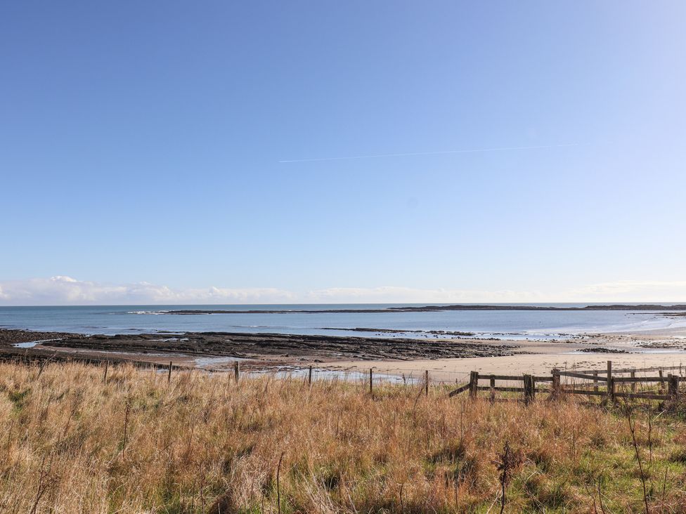 A coastal scene with grassy foreground, a wooden fence, rocky shoreline and calm sea at St Marys Cottages No1 in Low Newton-by-the-Sea