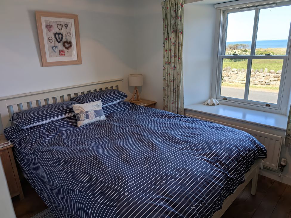 A bedroom with a bed with striped bedding bedside tables a lamp a framed artwork a window with a view of the sea and countryside at Beachward in Embleton