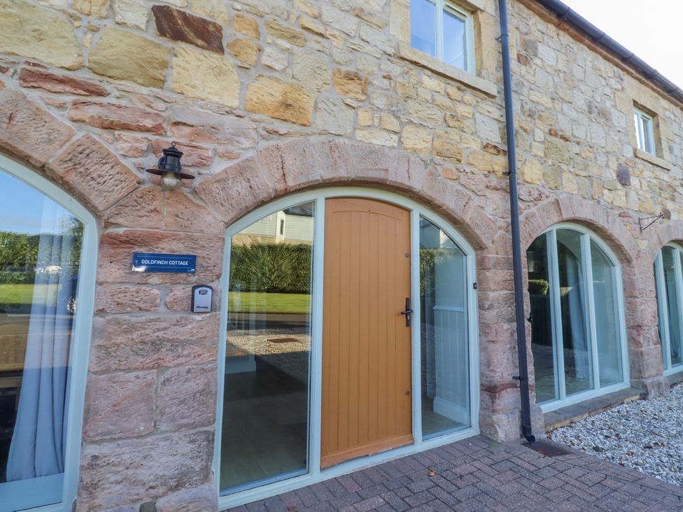 An exterior view of Goldfinch Cottage with a stone wall and an orange front door at Goldfinch in Lucker