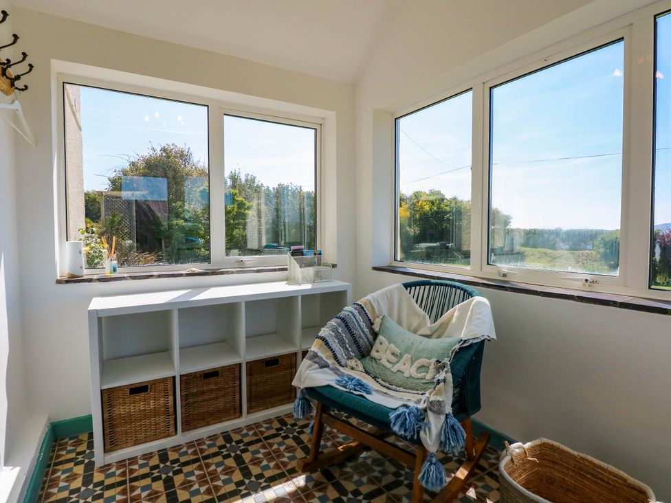 A sunroom with large windows a chair with a blanket and pillow a white shelving unit with baskets and a woven basket on the floor at Ael Y Fron in Aberdaron