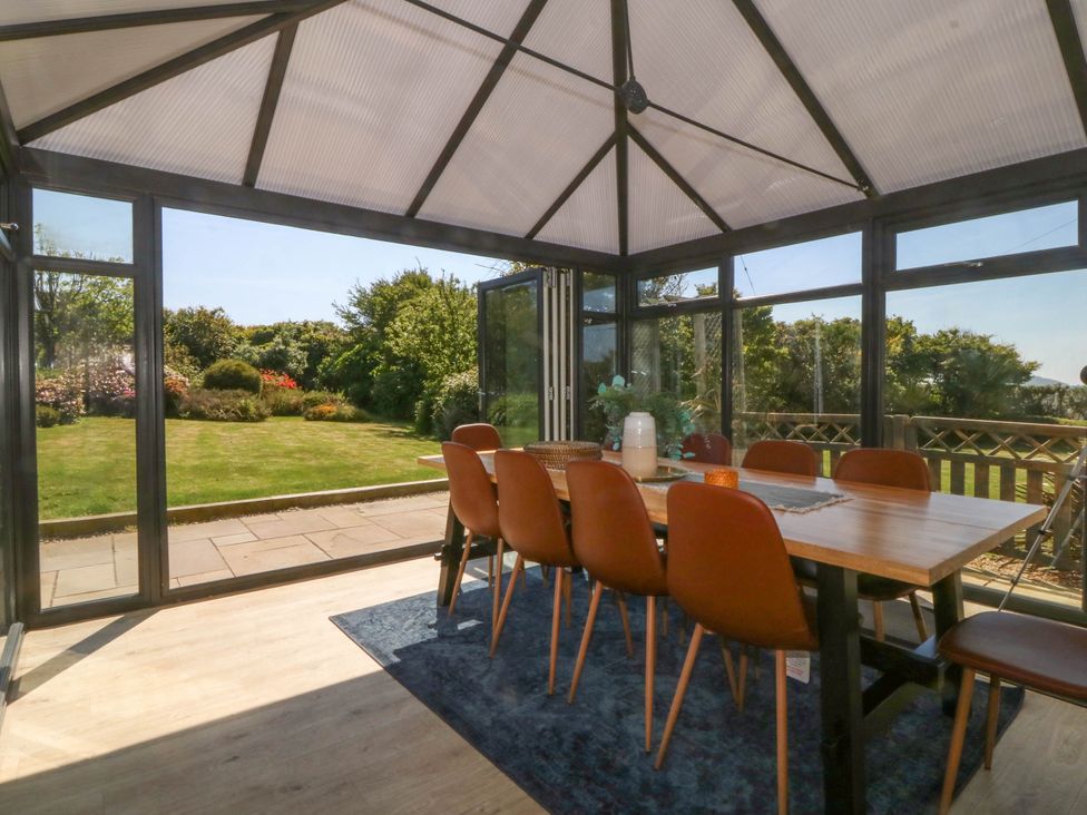 A dining area with a wooden table and brown chairs in a glass conservatory overlooking a garden at Ael Y Fron in Aberdaron