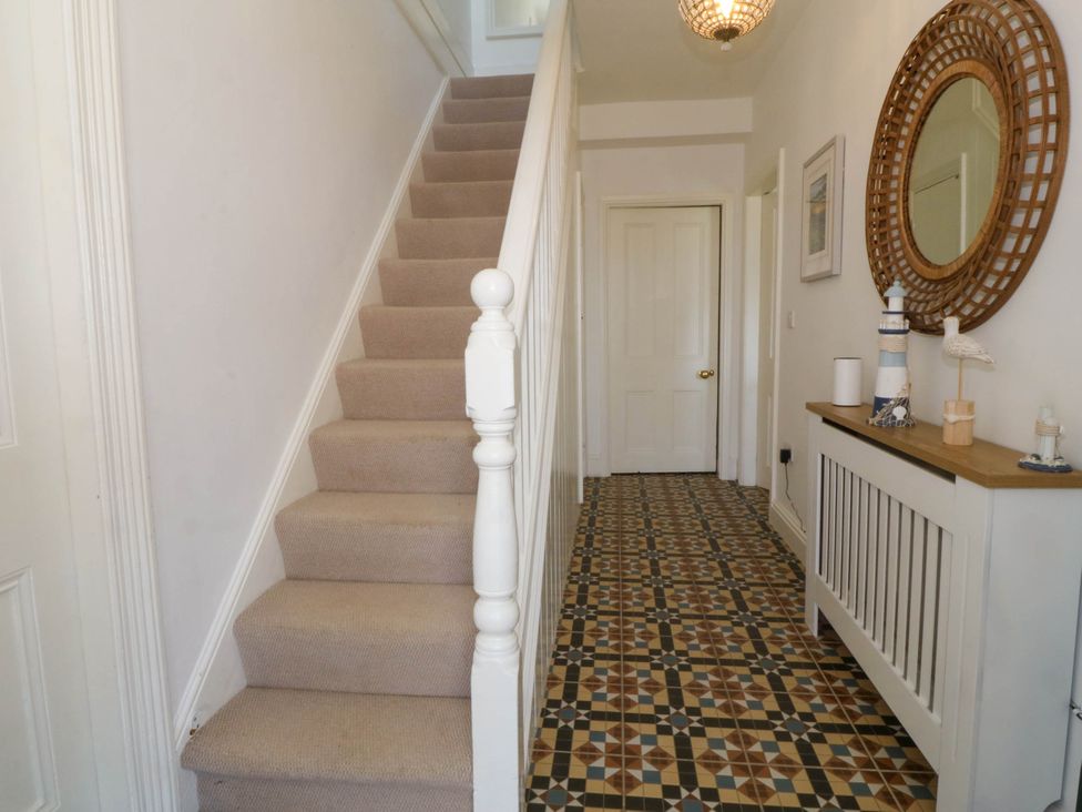 A hallway with carpeted stairs on the left mosaic tiled floor on the right side with a white radiator cover and decorative items with a round mirror above at Ael Y Fron in Aberdaron