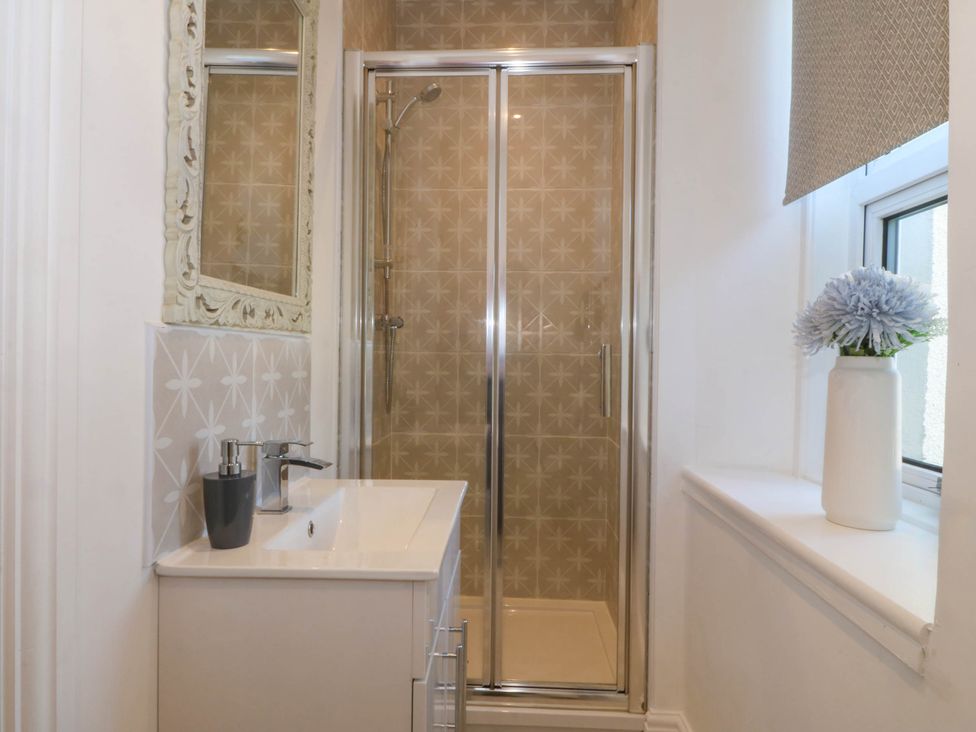 A small bathroom with a shower enclosure a sink with chrome faucet and soap dispenser a window with a vase of flowers and a patterned blind at Ael Y Fron in Aberdaron
