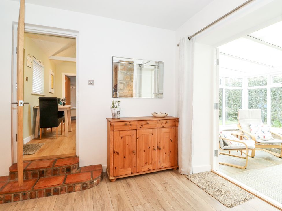 A hallway with wooden cabinet and mirror leading to a room with dining table and a sunroom with chairs and glass walls at Canny Cottage Low Hauxley Nr Amble