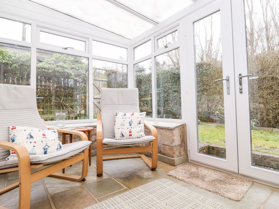 A conservatory with two wooden armchairs and cushions next to glass doors and windows showing a garden at Canny Cottage Low Hauxley Nr Amble