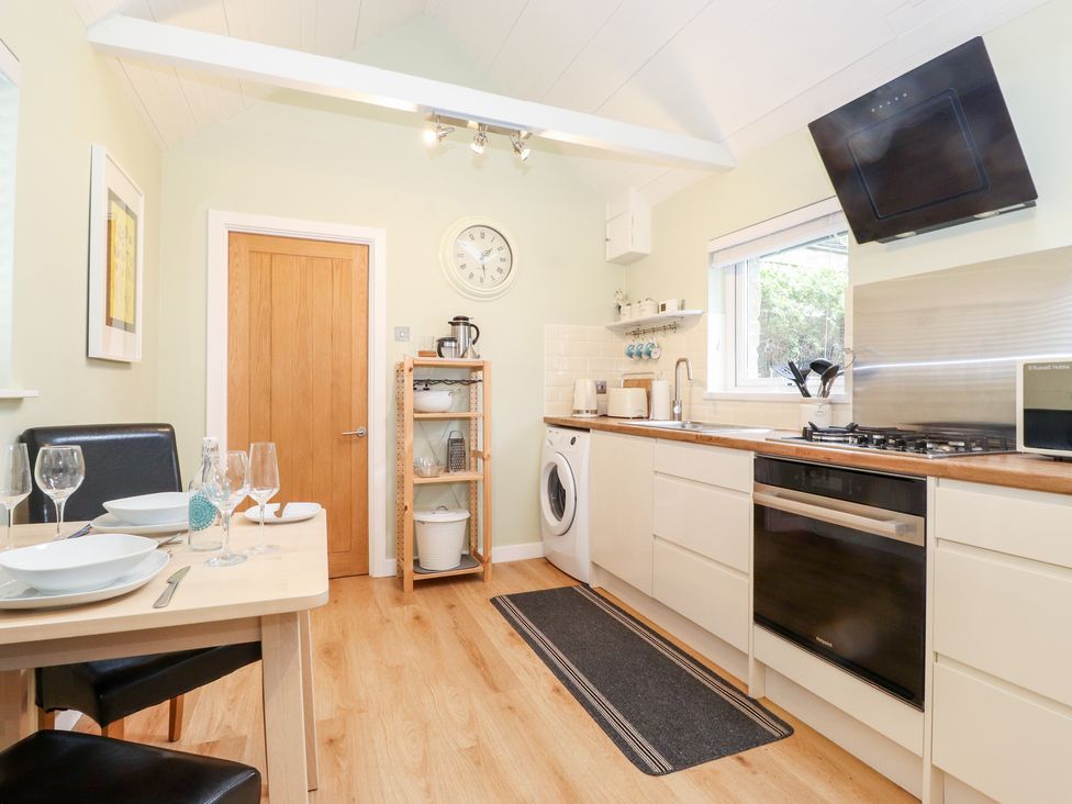 A kitchen with a dining table set with plates and glasses stove oven washing machine and wooden shelving at Canny Cottage Low Hauxley Nr Amble