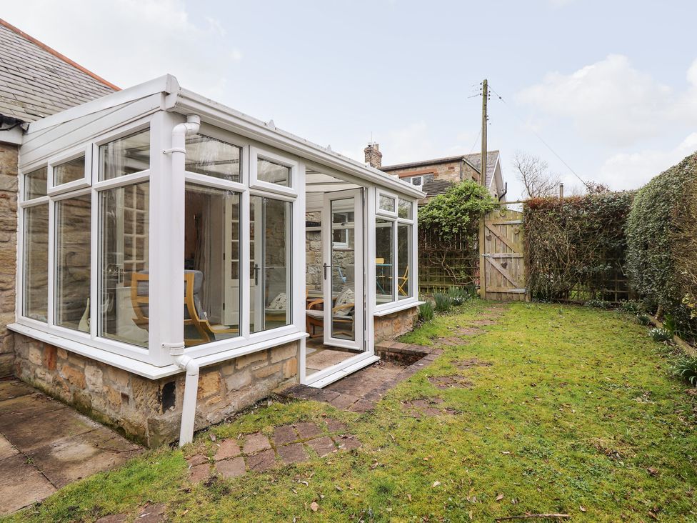 A glass conservatory attached to a stone house with a garden and wooden gate at Canny Cottage in Low Hauxley Nr Amble