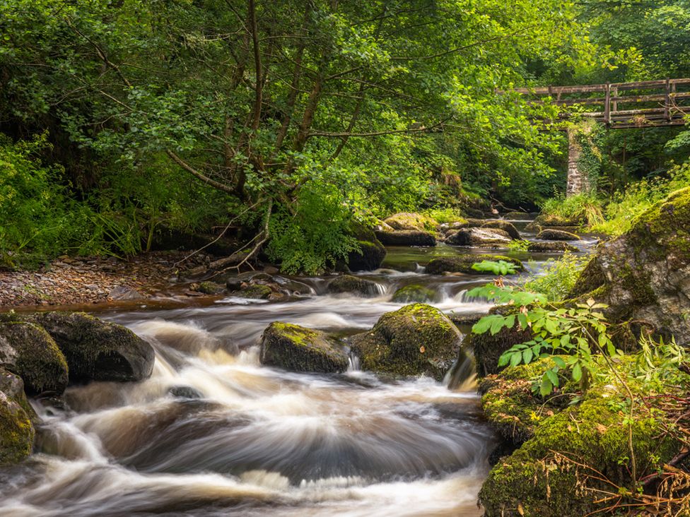 A river flowing over rocks with trees and a bridge at Larchwood Lodge @ Manleigh Park, Combe Martin