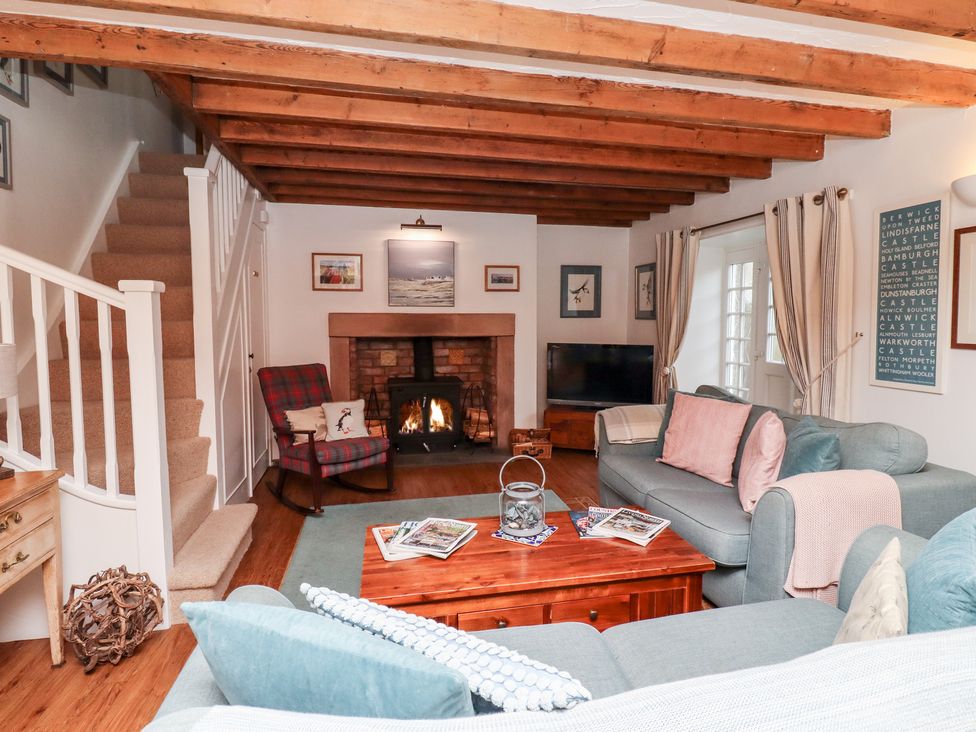 A living room with a wood beam ceiling a fireplace a plaid rocking chair a blue sofa a wooden coffee table and stairs at Seahares in Seahouses