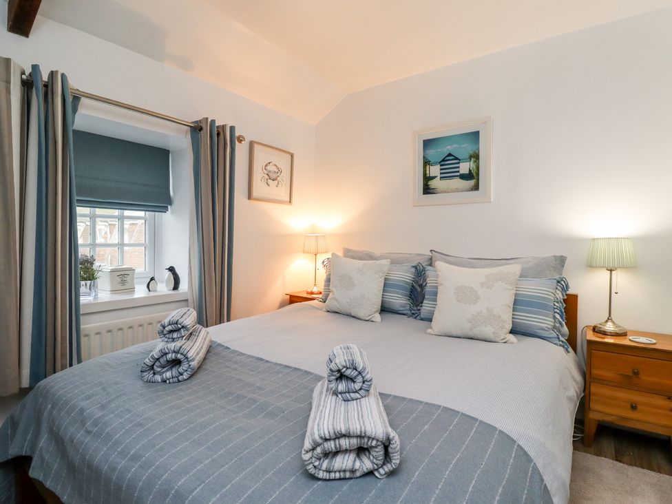 A bedroom with a bed with pillows and rolled towels two wooden bedside tables with lamps a window with curtains and a blind and framed pictures on walls at Seahares in Seahouses
