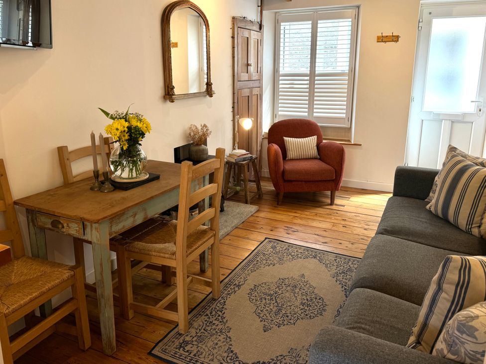 A living room with a wooden dining table and chairs a gray sofa with striped cushions a red armchair a rug a mirror on the wall and a window with shutters at Bodillys Cottage in Newlyn