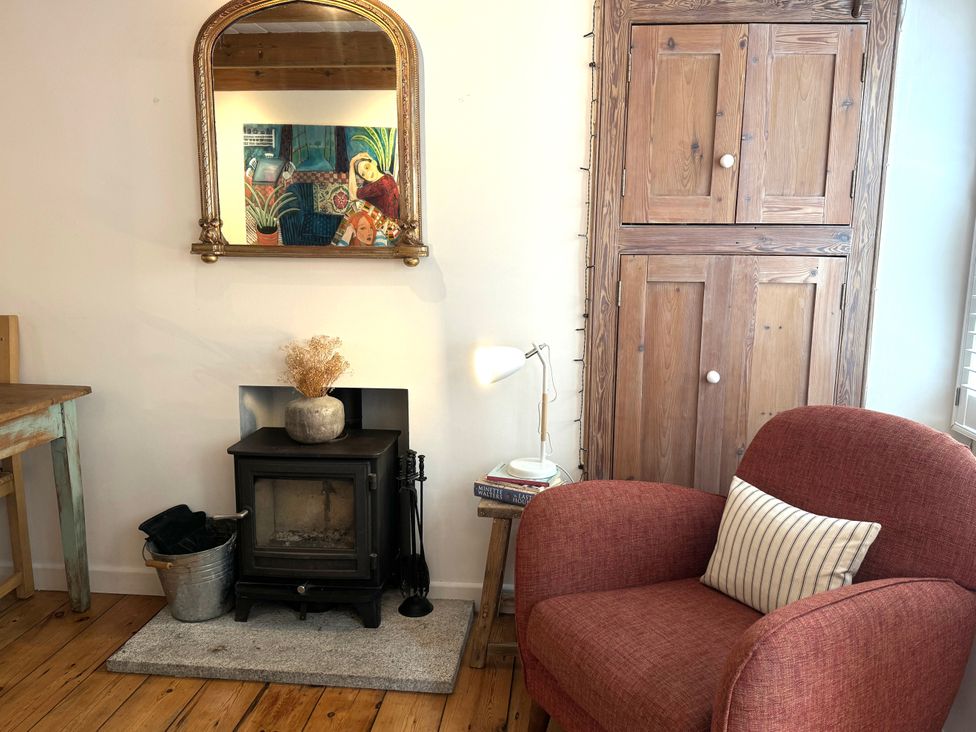 A room with a red armchair next to a wooden cabinet and a small stove with a vase on top at Bodillys Cottage in Newlyn