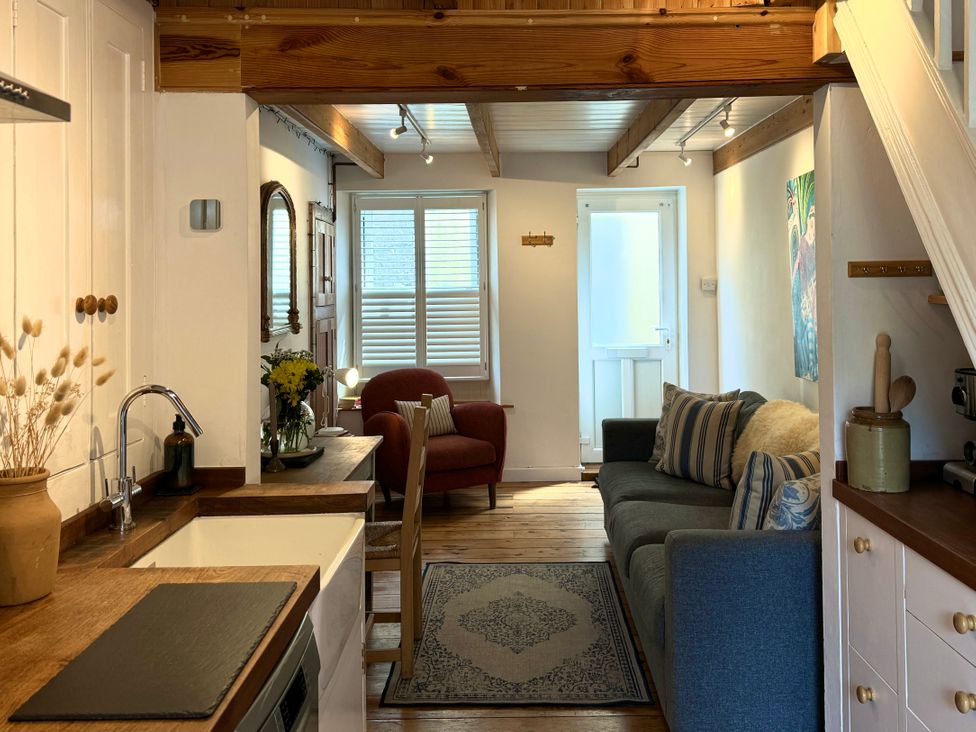 A small living area with a kitchen sink counter a sofa with cushions an armchair a wooden chair a rug wooden beams on the ceiling and a door with shutters at Bodillys Cottage in Newlyn