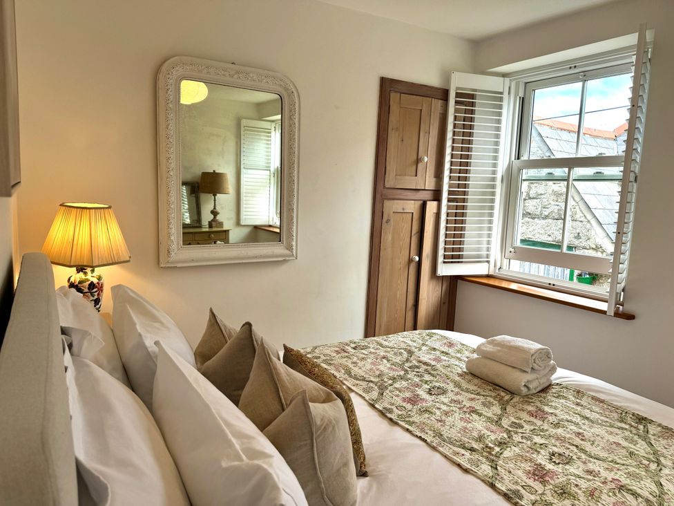 A bedroom with a bed with pillows and folded towels on a patterned throw near a window with shutters and a wall mirror at Bodillys Cottage in Newlyn