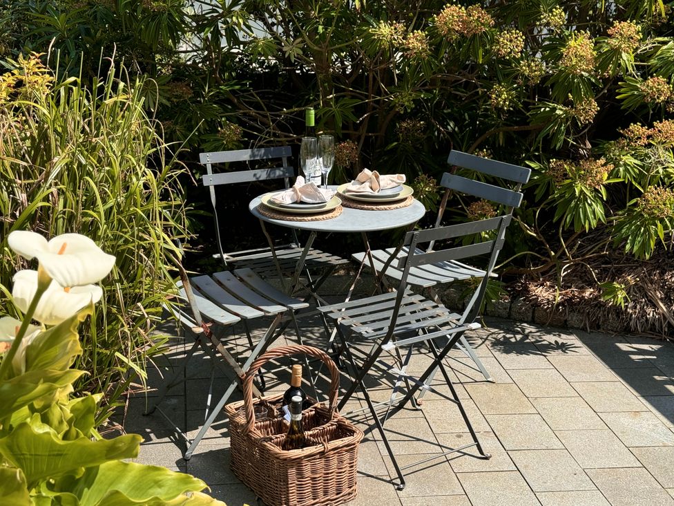 An outdoor patio with a round table set for two with plates and glasses surrounded by four metal chairs and a wicker basket with bottles of wine at Bodillys Cottage in Newlyn