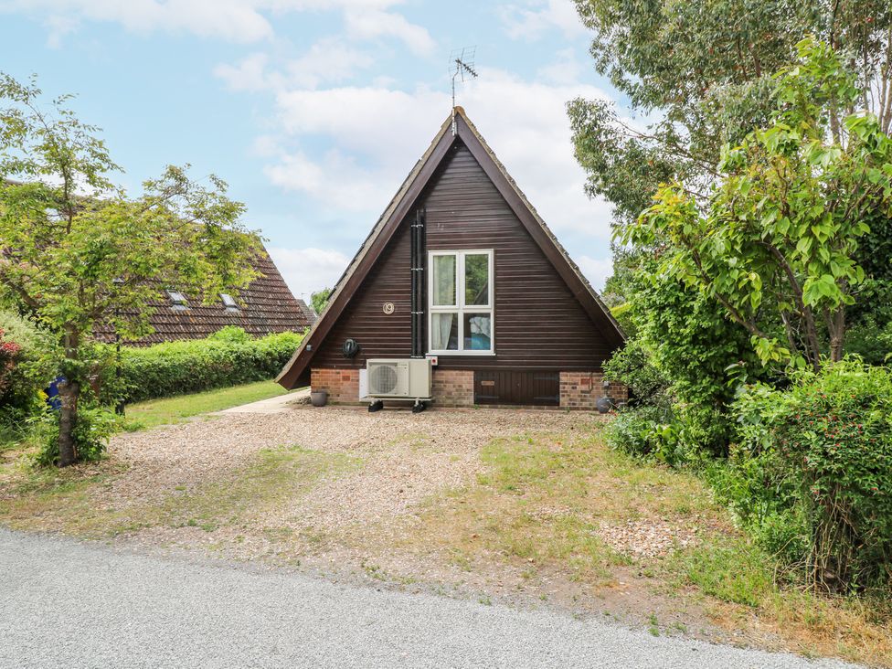 An A-frame house with wooden siding and a gravel driveway surrounded by trees and bushes at Kingfisher in Isleham
