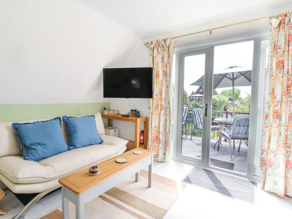 A living room with a cream sofa with blue cushions a wooden coffee table a wall mounted tv floral curtains and glass doors leading to a patio at Kingfisher in Isleham