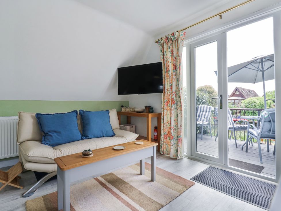 A living room with a sofa, coffee table, wall-mounted TV, floral curtains, and glass doors opening to an outdoor balcony at Kingfisher in Isleham