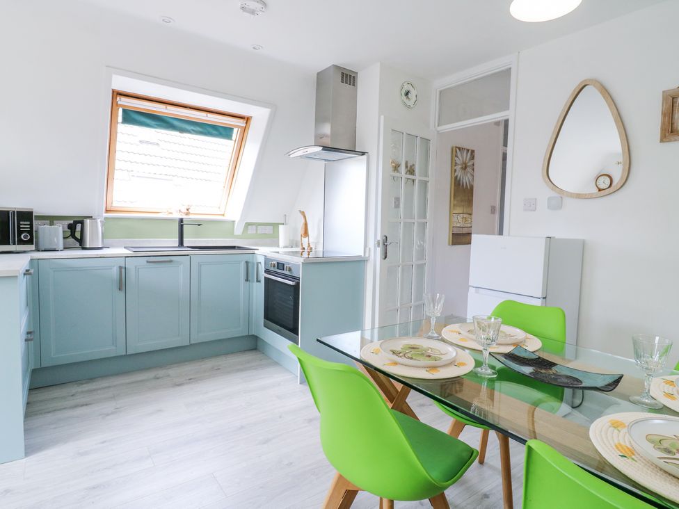 A kitchen with blue cabinets green chairs glass dining table and a window at Kingfisher in Isleham