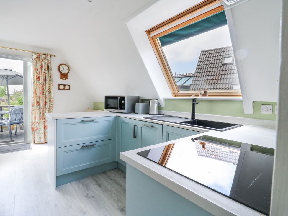 A kitchen with blue cabinets a black induction stove a microwave and a large window overlooking rooftops at Kingfisher in Isleham