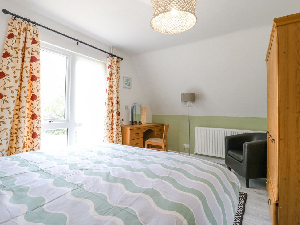 A bedroom with a bed and patterned curtains next to a window a wooden desk with a chair a wardrobe a black armchair and a floor lamp at Kingfisher in Isleham