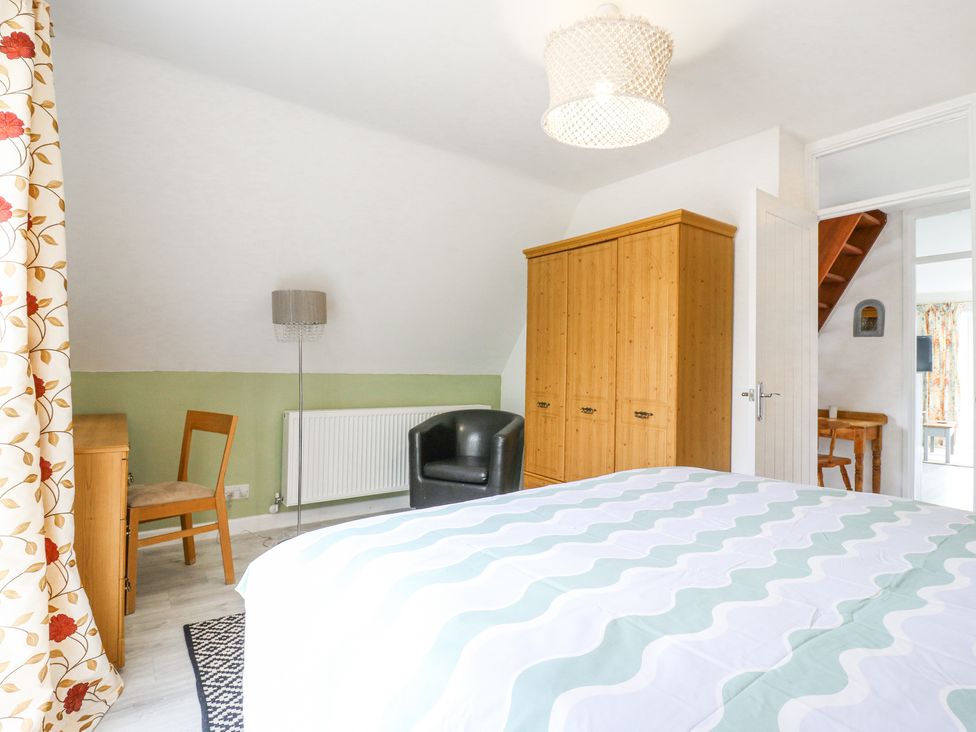 A bedroom with a wooden wardrobe a black armchair a wooden chair and desk patterned curtains and a bed with a patterned bedspread at Kingfisher in Isleham