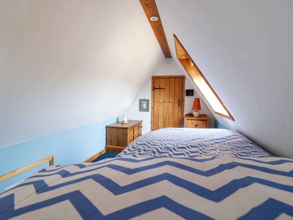 A small attic bedroom with a bed covered in blue and white zigzag patterned bedding two wooden dressers a slanted ceiling with a skylight and a wooden door at Kingfisher in Isleham