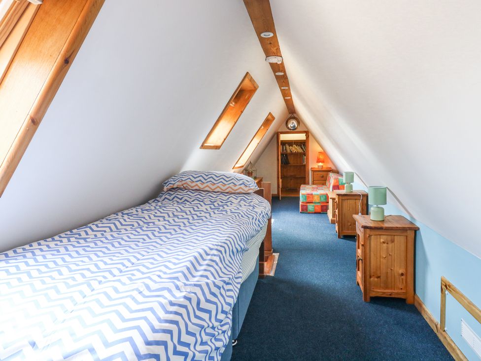 A narrow attic bedroom with a single bed a chevron patterned duvet and wooden furniture including bedside tables and a bookshelf at Kingfisher in Isleham