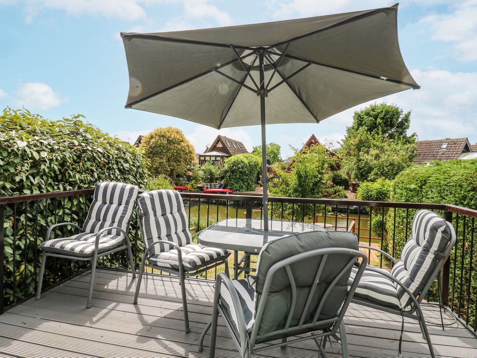 An outdoor deck with a table an umbrella and four cushioned chairs overlooking greenery and houses at Kingfisher in Isleham