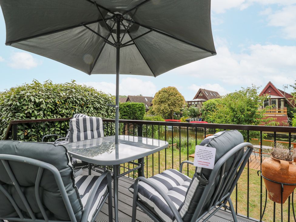 A balcony with a glass table and four cushioned chairs under an umbrella overlooking a garden with houses and greenery at Kingfisher in Isleham
