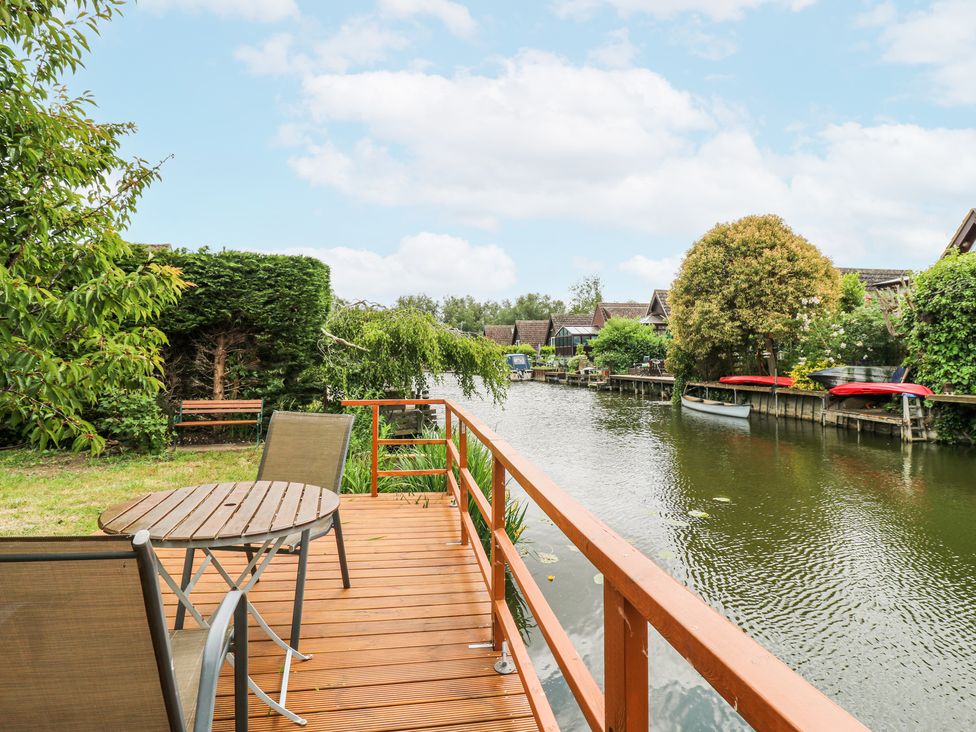 A wooden deck with table and chairs overlooking a canal with boats and houses at Kingfisher Isleham