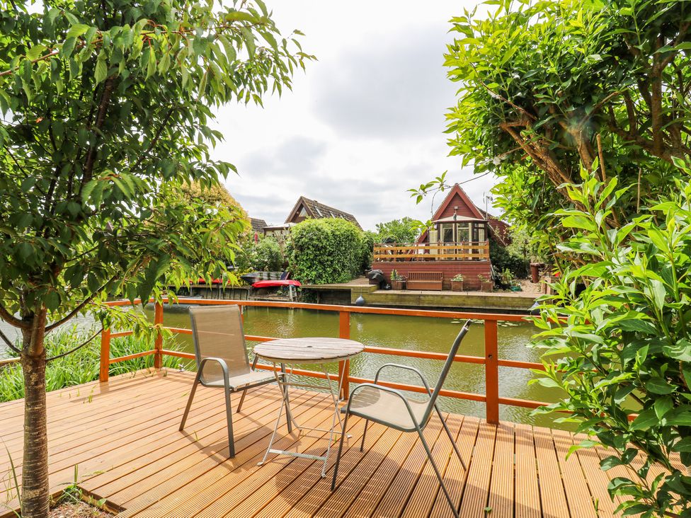A wooden deck with two chairs and a round table overlooking a water canal with houses and greenery at Kingfisher in Isleham