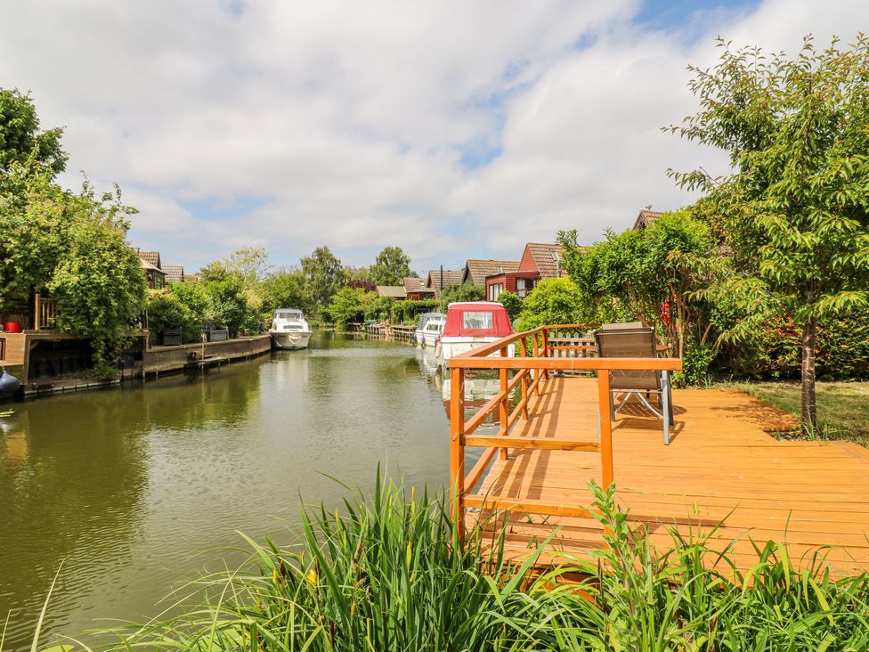 A wooden deck with chairs by a river with boats and houses along the riverbank at Kingfisher in Isleham