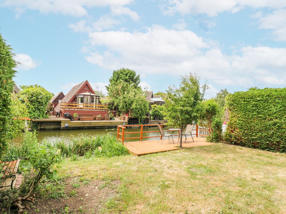 A garden with a wooden deck and chairs beside a river with houses on the opposite bank at Kingfisher in Isleham