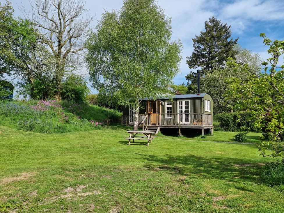 A small house with a wooden porch surrounded by trees and grass with a picnic table on the lawn at Mucklewick Hollow in Minsterley near Bishops Castle