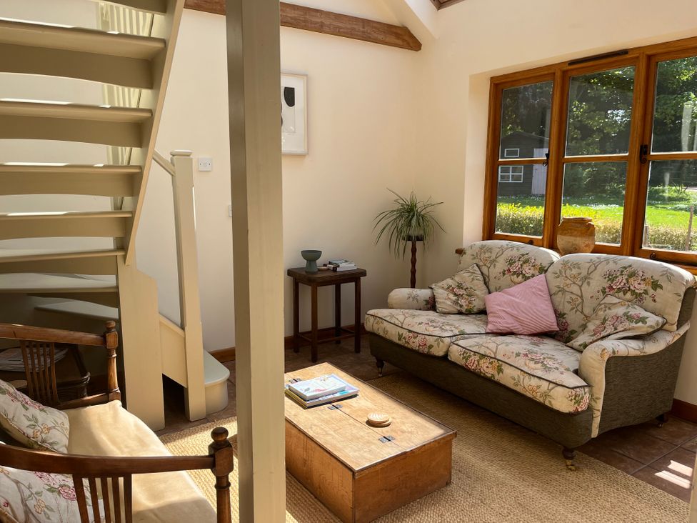 A living room with floral sofas a wooden coffee table a wooden armchair a side table with books a plant and a staircase by a large window at The Apple House in Spaxton near Nether Stowey
