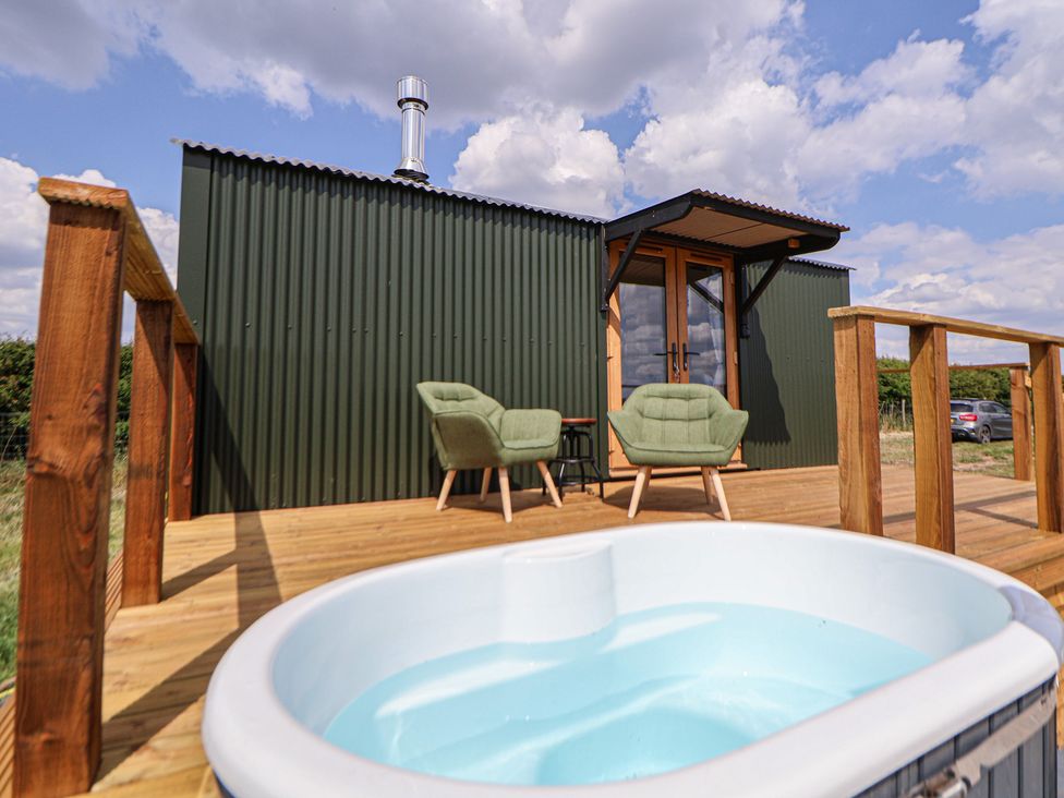 A wooden deck with green chairs and a small table outside a green corrugated metal building with a hot tub in the foreground at The Coop in Aston near Woore