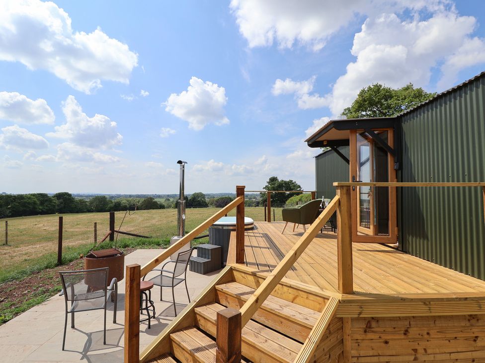 An outdoor wooden deck with stairs chairs a table and a hot tub overlooking a grassy field at The Coop in Aston near Woore