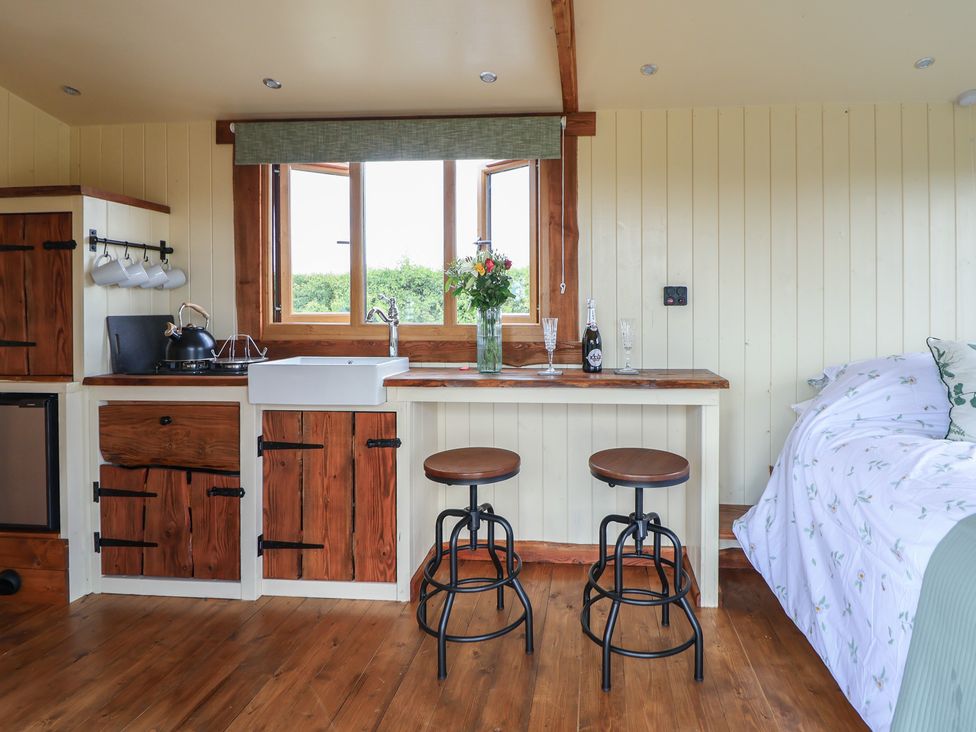 A small kitchen with wooden cabinets a sink two stools a bottle and glasses by a window and a bed with floral bedding at The Coop in Aston near Woore