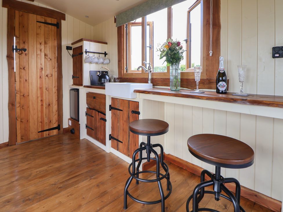 A kitchen area with wooden stools a sink flowers on the counter and a bottle with glasses at The Coop in Aston near Woore