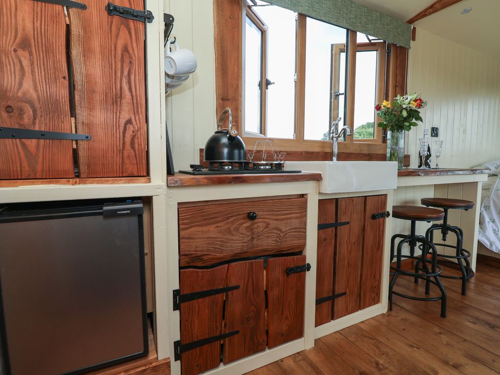 A kitchen area with wooden cabinets a small fridge a black kettle a white sink two stools and flowers on the counter at The Coop in Aston near Woore