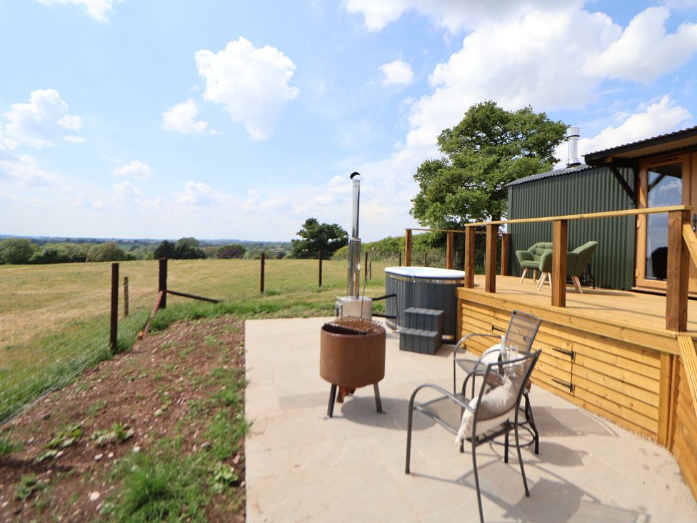 An outdoor patio with chairs a fire pit a hot tub and a wooden deck next to a green building in a rural area at The Coop in Aston near Woore