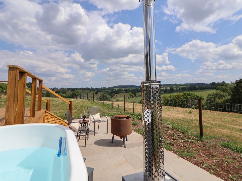 An outdoor patio with a hot tub chairs a small table and a fire pit overlooking green fields at The Coop in Aston near Woore